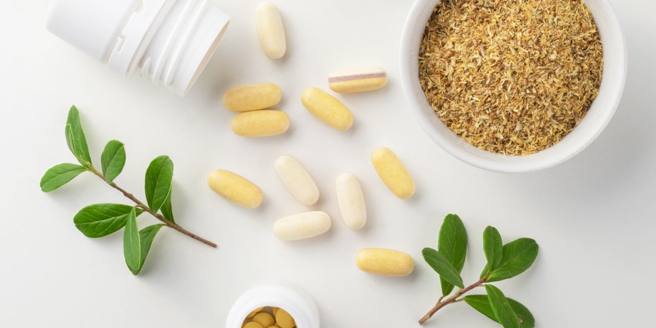 herbal supplement capsules with green leaves and a bowl of dried plant material on a white surface