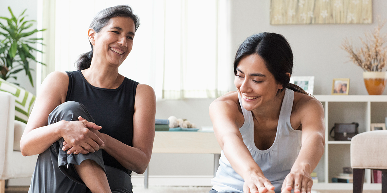 Two people in workout clothes stretch on a living room floor with shelves and plants in the background.