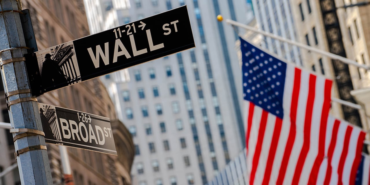 Close up photo of the street signs at the corner of Wall Street and Broad Street with American flags and buildings in the background. 